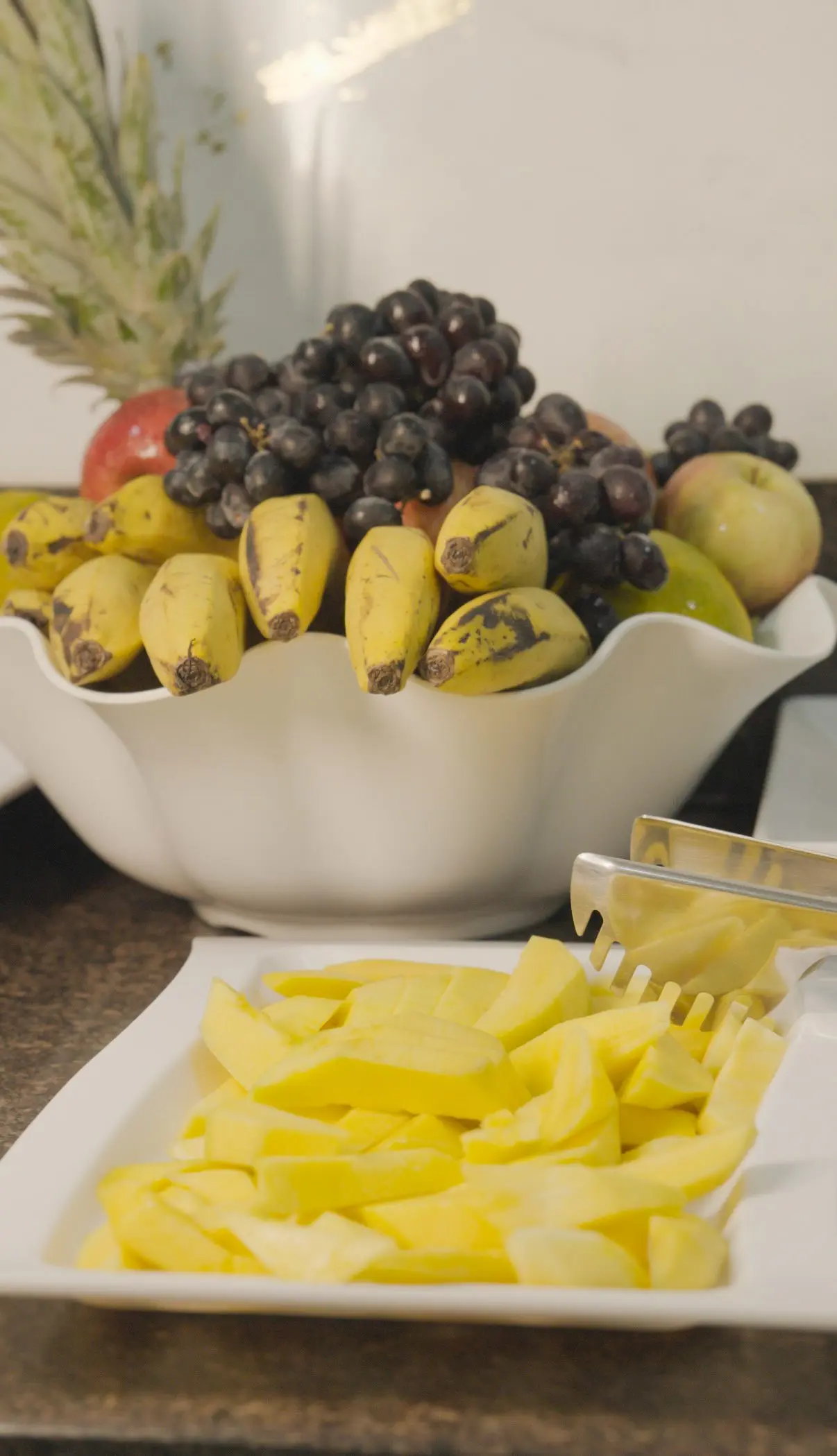 Fruteira com uvas, bananas e maçãs e travessa de abacaxi fatiado no café da manhã