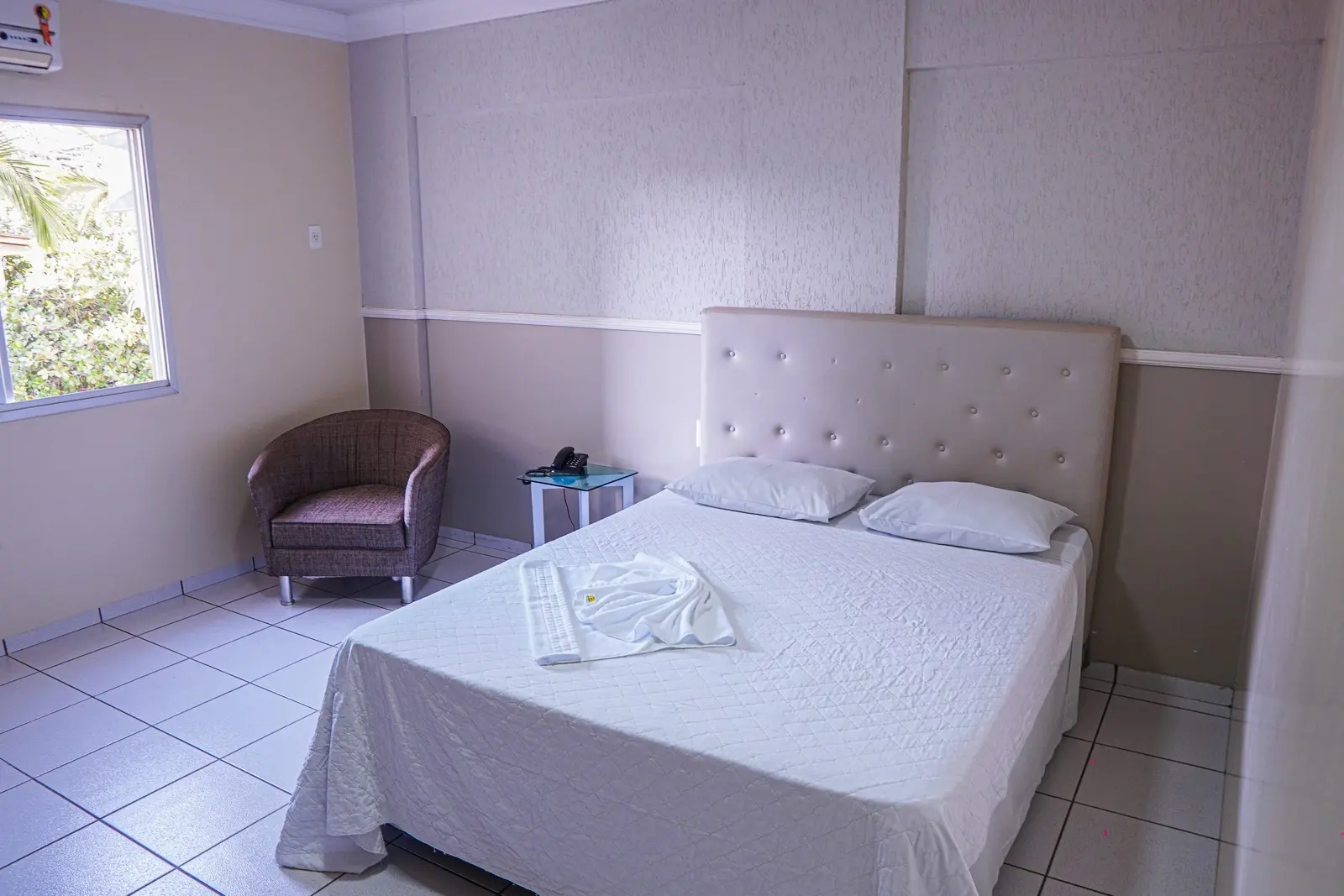 Quarto Individual com cama de casal arrumada, poltrona e janela