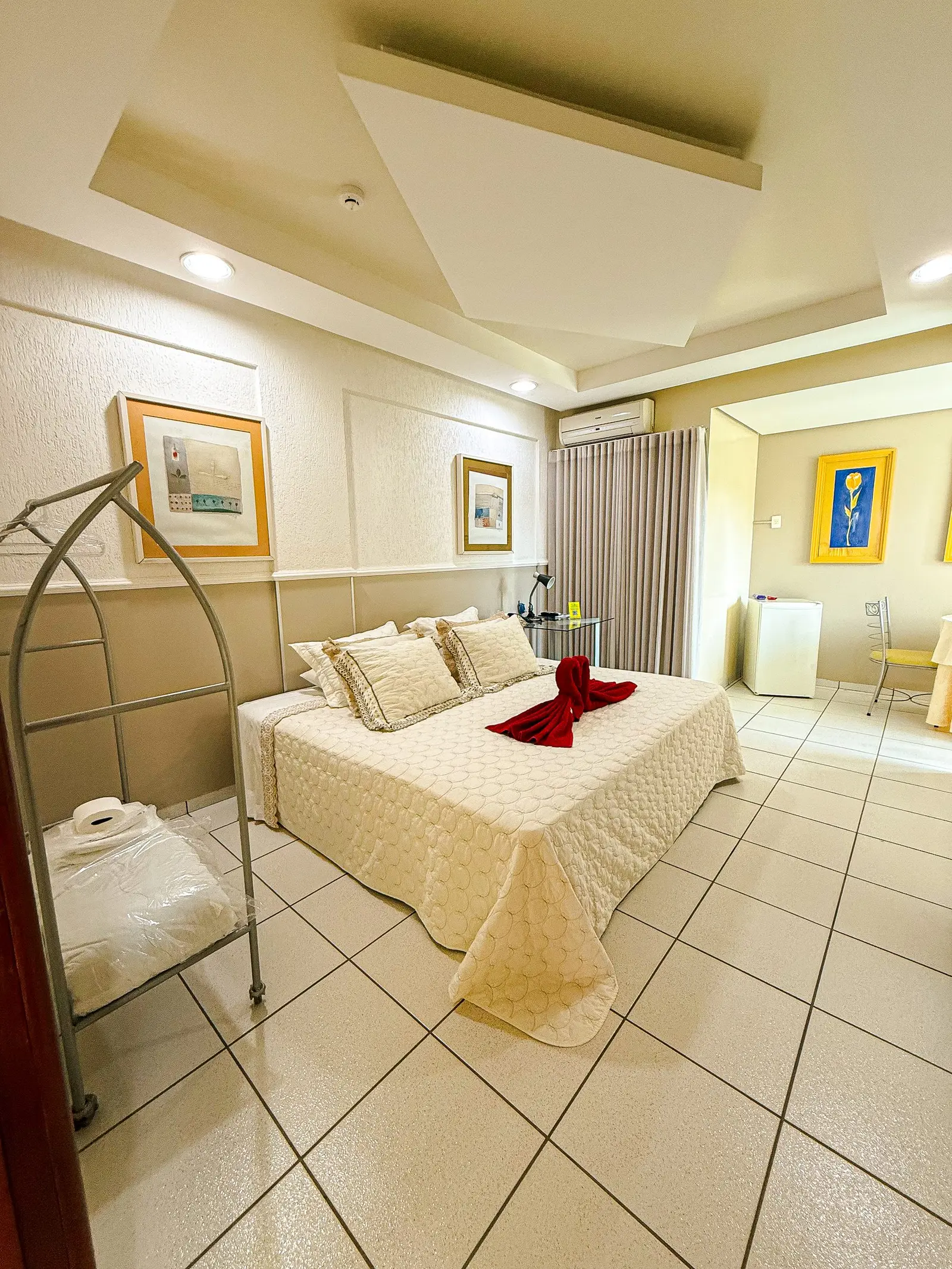 Quarto com cama de casal, toalha vermelha sobre a cama e carrinho de bagagem